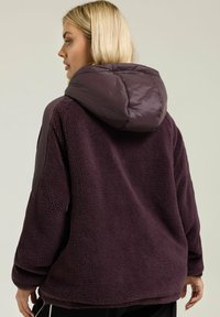 Fioletowa polarowa kurtka z pikowaną, błyszczącą kapturem; charakteryzuje się luźnym krojem i teksturowanym materiałem; zawiera boczne kieszenie i zaokrąglony dół.