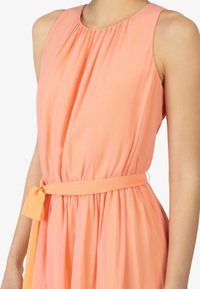 Perzikkleurige mouwloze jurk met een verzamelde halslijn, een tailleband en vloeiende stof, met een zachte, gladde textuur.