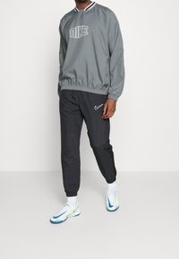 Pullover grigio con logo "Nike" bianco, vita elastica e polsini; abbinato a pantaloni jogger neri con il baffo bianco "Nike". Scarpe sportive bianche e blu.