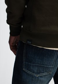 Donkergroene sweatshirt met ribbels aan de mouwen, voorzien van een geweven label, gecombineerd met blauwe denim jeans met een detail aan de achterzak.