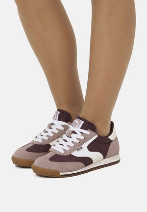 Person, die mauve- und weinrote Sneakers mit weißen Schnürsenkeln und Gummisohlen trägt und auf einer weißen Fläche steht.