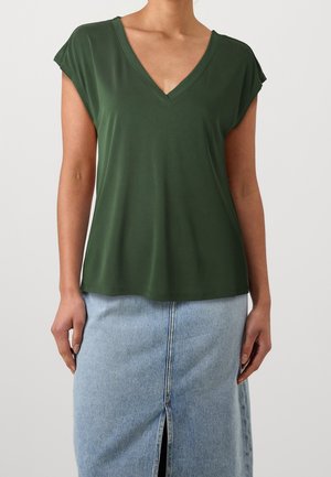 Femme portant un haut vert foncé à manches courtes et col en V et une jupe en jean bleu clair avec une fente à l'avant sur un fond uni.