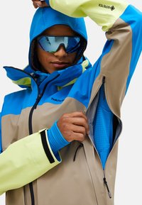 Outdoorjacke mit blauem oberen Teil und beigem unteren Teil. Verfügt über einen hellgelben Ärmel, Reißverschlüsse und eine Kapuze. Texturiertes Innenfutter sichtbar.