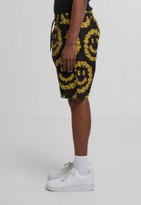 Sorte shorts med gult blomster-smilfjesmønster, elastisk midjebånd, snøring og sidelommer, kombinert med hvite sneakers og sokker.