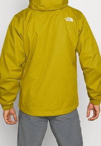 Chaqueta amarilla impermeable con capucha, puños elásticos y un logo blanco en la parte trasera. Textura suave, ajuste casual. Combinada con pantalones grises.