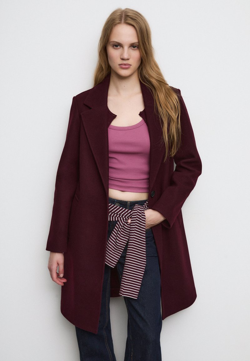 Burgunderfarbener Wollmantel mit Reverskragen, einem Knopf und Seitentaschen, über einem pinken Tanktop und dunklen Jeans mit einem gestreiften Gürtel getragen.