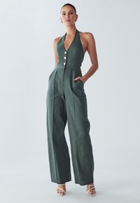 Zelený overal se zavazováním za krk, s přiléhavým živůtkem, zlatými knoflíkovými detaily, nohavicemi širokého střihu a dvěma bočními kapsami, vyrobený z texturované látky.