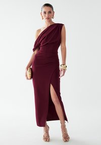 Burgundy šaty na jedno rameno s drapovaným telom, priliehavým pásom a rozparkom až po stehno. Kombinované so zlatými šperkami a sandálmi na remienky.