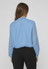 Blusa blu chiaro con un motivo a zigzag testurizzato, maniche lunghe, colletto volant e chiusura con bottone a oblò sul retro.