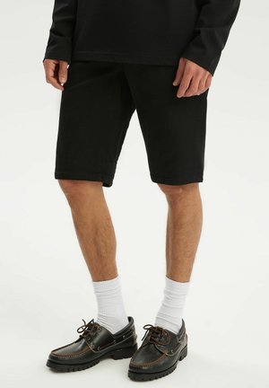 Męskie nogi ubrane w czarne spodenki, białe skarpety sportowe i czarne buty żeglarskie z brązowymi sznurowadłami na jasnym tle.