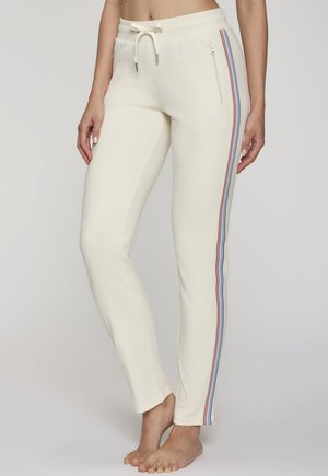 Schmal geschnittene cremefarbene Jogginghose mit roten, blauen und schwarzen Streifen an der Seite, Reißverschlusstasche und Kordelzugbund, getragen von einem barfuß stehenden Modell.