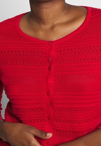 Cardigan rosso lavorato a maglia con chiusura anteriore a bottoni, caratterizzato da strisce orizzontali e motivi traforati. Tessuto morbido e design aderente.