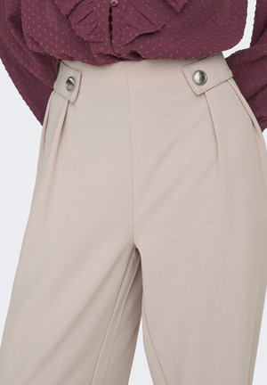 Pantalon plissé beige à texture lisse, doté de boutons argentés à la taille, associé à un chemisier bordeaux à motifs.