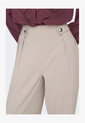 Pantalon plissé beige à texture lisse, doté de boutons argentés à la taille, associé à un chemisier bordeaux à motifs.