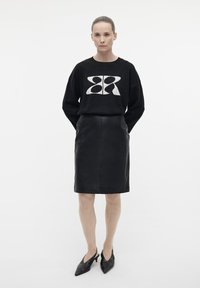 Zwarte oversized trui met een crème abstract patroon, gecombineerd met een aansluitende zwarte leren rokje. Model draagt zwarte puntige hakken.