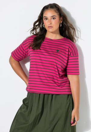 Frau mit langen dunklen Haaren trägt ein pink-schwarz gestreiftes T-Shirt und einen grünen Rock, steht mit einer Hand in der Hüfte vor einer weißen Wand.