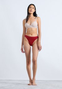 Nude gewatteerde bh met verstelbare bandjes en rode high-waist slipjes, beide van gladde stof, gepresenteerd op een model tegen een lichte achtergrond.