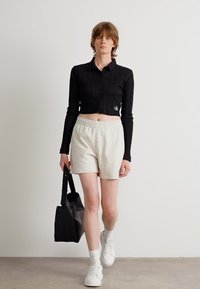 Svart cropped skjorte med knapper, lys grå shorts, hvite joggesko og en svart tote bag. Glatte teksturer og minimalistisk design.