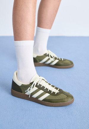Nogi w białych skarpetach do kostek i oliwkowozielonych butach Adidas z białymi paskami, stojące na jasnoniebieskim dywanie.