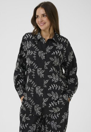 Woman smiling, wearing a black long-sleeve buttoned shirt and matching pants with white leaf and vine embroidery pattern.