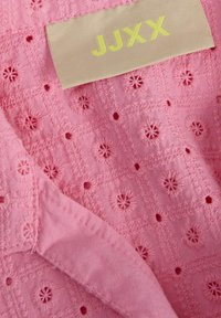 Roze tkanina s izvezenim cvjetnim uzorcima i malim rupama, s bež oznakom sa žutim natpisom "JJXX" u gornjem lijevom kutu.