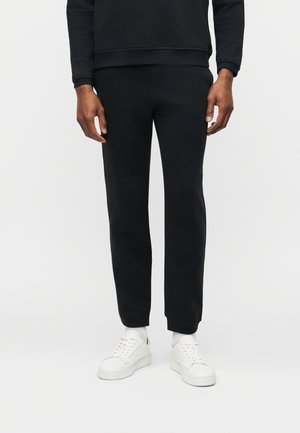 Man die zwarte joggingbroek, zwarte trui, witte sokken en witte sneakers draagt, staand tegen een effen witte achtergrond.