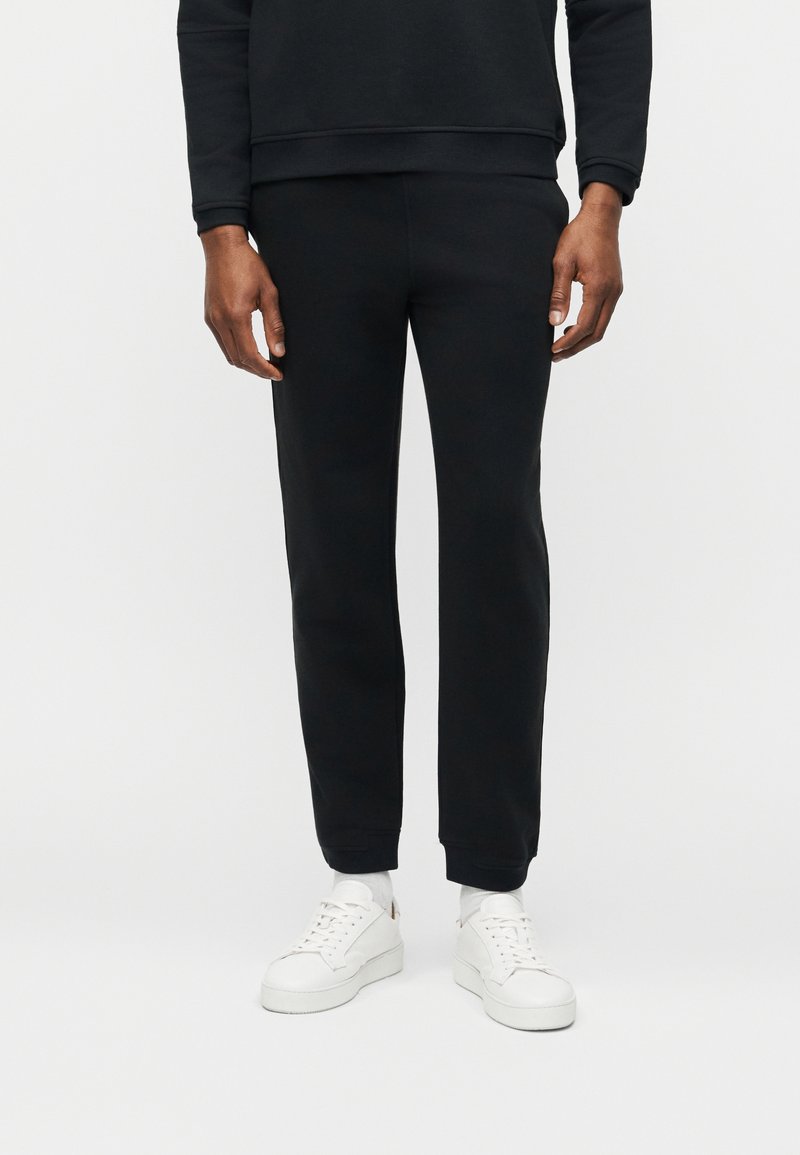 Άνδρας που φοράει μαύρο παντελόνι φόρμας, μαύρο φούτερ, λευκές κάλτσες και λευκά αθλητικά παπούτσια, στέκεται μπροστά σε λευκό φόντο.