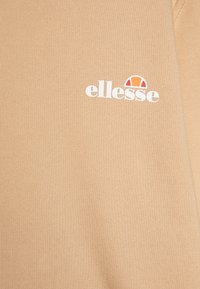 Bež dukserica s rebrastom teksturom; sadrži bijeli "ellesse" logo i crveni i narančasti naglasak iznad natpisa.
