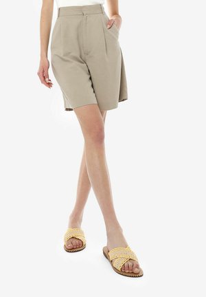 Pantalones cortos beige de talle alto con pliegues delanteros, de longitud hasta la rodilla, combinados con sandalias de tiras trenzadas amarillas. Textura de tela suave, diseño casual.