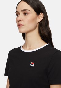 Zwarte katoenen T-shirt met korte mouwen, witte kraagafwerking en een vierkant geborduurd logo in rood, wit en blauw op de borst.