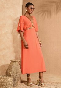 Koraal maxi jurk met een V-hals en paillettenaccenten op de mouwen en taille. G gedragen met gouden wedge sandalen en oversized zonnebril.