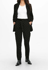 Woman wearing black blazer and pants with white polka dots, white t-shirt, with hands in pockets and black ankle boots.