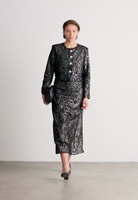 Moteris eina juodu ir sidabriniu raštuotu ilgu sijonu ir atitinkamu ilgu rankoviu švarku, laiko juodą rankinėje.