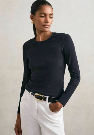 Tumehadega naine musta pikakarvalise särgiga, valgete pükste, musta kuldse nööbiga vööga ja kuldsete kõrvarõngastega, seisab üks käsi taskus.