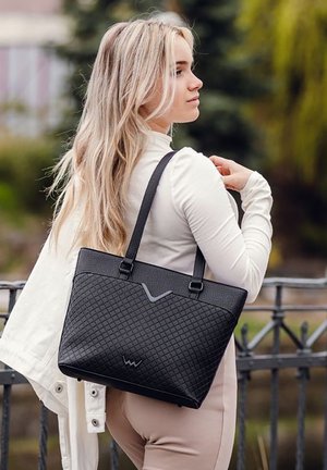 Schwarze Tragetasche mit einem strukturierten Diamantmuster, das kontrastierende graue Akzente und doppelte Schulterriemen aufweist, angelehnt an ein hellfarbenes Outfit.