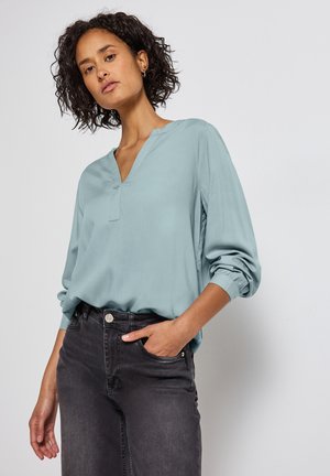 Blouse bleu clair à manches longues avec un décolleté en V et des poignets élastiques. Associée à un jean en denim gris foncé, comportant une poche avant et un détail de bouton.