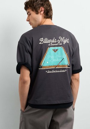 Young man wearing dark t-shirt with rolled sleeves and billiards table graphic and text "Billiards Night at Basement Club" on the back.
