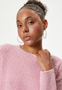Pull en texte rose avec un col rond et des manches longues. Le modèle porte des boucles d'oreilles en créole, a la peau lisse et les cheveux bouclés tirés en arrière.