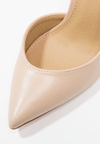Chaussure en cuir beige à bout pointu avec une texture lisse et des coutures visibles le long du bord, présentée sur un fond blanc.