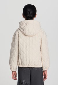 Giacca beige trapuntata con cappuccio, caratterizzata da un motivo a diamante e delicati accenti brillanti. I polsini e l'orlo elastico garantiscono una vestibilità aderente.