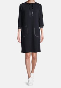 Czarna sukienka dresowa z trzykwartowymi rękawami, dwiema przednimi kieszeniami, satynowymi trokami i błyszczącymi akcentami na rękawach. Połączona z białymi sneakerami.