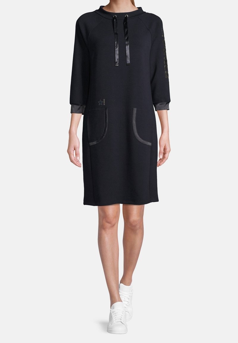 Czarna sukienka dresowa z trzykwartowymi rękawami, dwiema przednimi kieszeniami, satynowymi trokami i błyszczącymi akcentami na rękawach. Połączona z białymi sneakerami.