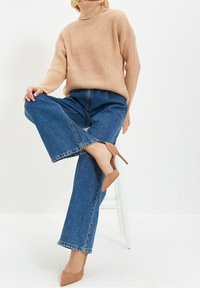 Jasnoróżowy sweter z golfem i prążkowaniem, niebieskie szerokie jeansy oraz beżowe buty na obcasie, a osoba siedzi na białym stołku.