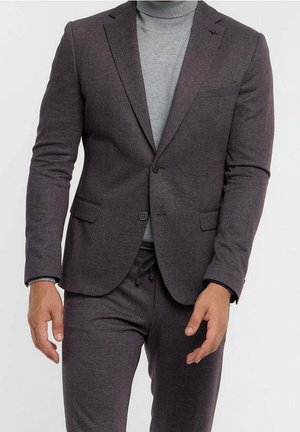 Mann trägt einen dunkelgrauen Blazer mit passenden Hosen und einen hellgrauen Rollkragenpullover vor einem schlichten Hintergrund.