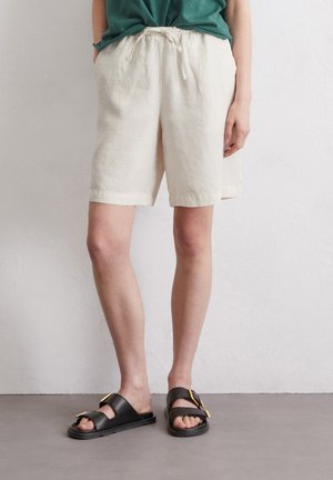 Shorts en lin de couleur crème avec une taille élastique et un cordon de serrage. Assortis à des sandales noires avec une accent en boucle dorée.