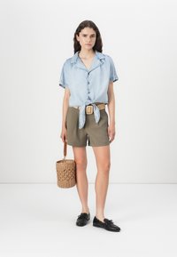 Ανοιχτό μπλε τζιν πουκάμισο κοντομάνικο με κόμπο μπροστά, συνδυασμένο με ελαιόχρωμες σορτς και ένα υφαντό στρογγυλό τσάντα. Μαύρα παπούτσια με κορδόνια ολοκληρώνουν την εμφάνιση.