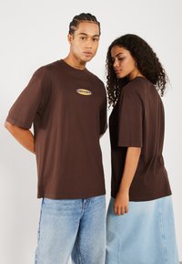 T-shirts marron surdimensionnés avec un col rond et des manches courtes. L'un d'eux affiche un logo sur la poitrine. Portés avec un jean en denim bleu clair et une jupe.