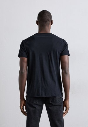 Mand stående med ryggen mod, iført en ensfarvet sort t-shirt og mørke jeans mod en lysegrå baggrund.
