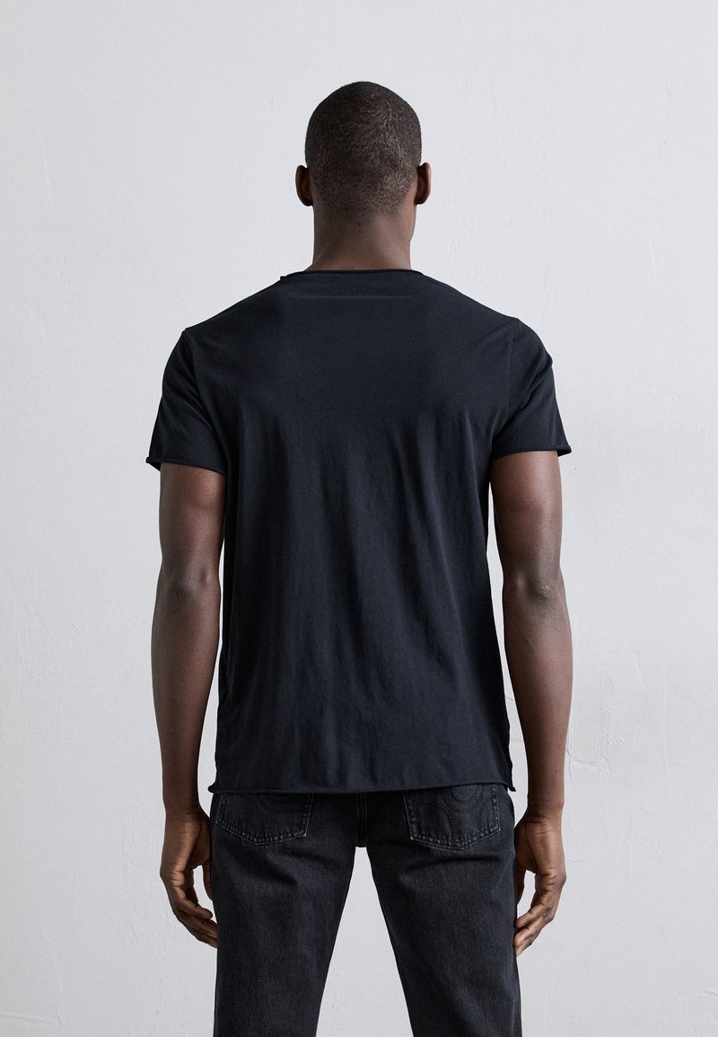 Homme debout de dos, portant un t-shirt noir uni et un jean foncé sur un fond gris clair.