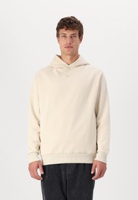 Sudadera de algodón crema con bolsillo canguro, tejido texturizado y puños acanalados, que presenta un pequeño logo en el pecho.